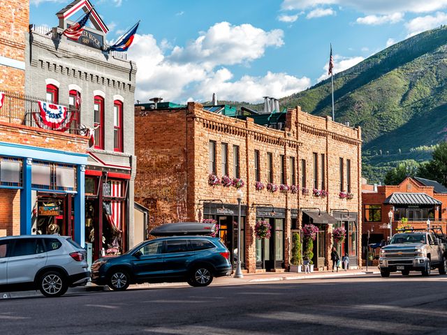 A street in Aspen on a sunny day in spring or summer with brick buildings and a few cars with green mountains in the background.