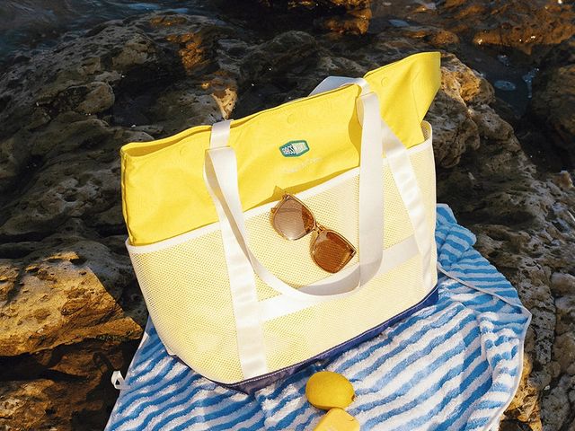 A yellow beach bag on a striped blue towel with sunglasses attached.
