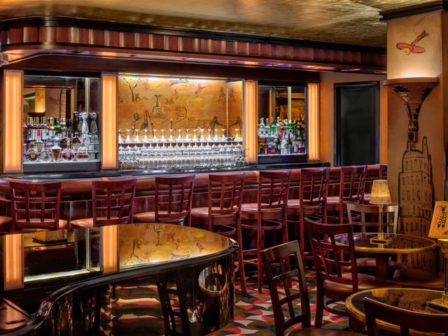 An empty hotel bar with red and gold accents, a painted mural behind the bar, and patterned carpeting.