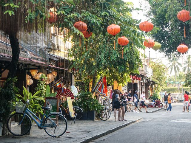 Several storefronts on a street in Hoi An, Vietnam with paper lanterns hanging and many green trees.