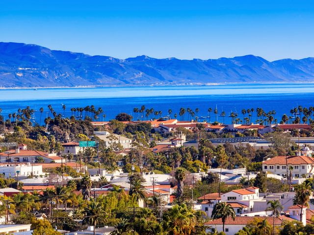 An aerial view over the coastline of Santa Barbara with buildings, a beach, and mountains in the distance.