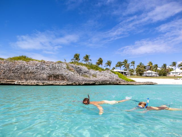 Two people seen snorkeling in the water in clear blue water next to a beach with small cliffs.