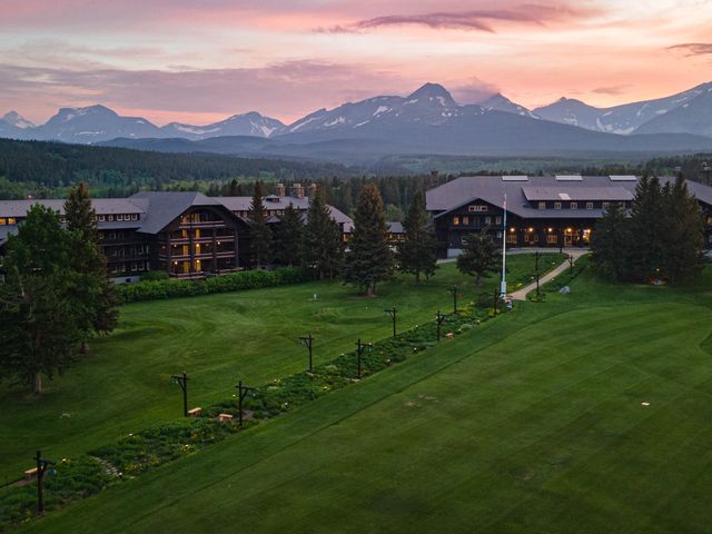 An aerial view of a large lodge hotel with green lawns and snow-capped mountains in the distance, at sunset.