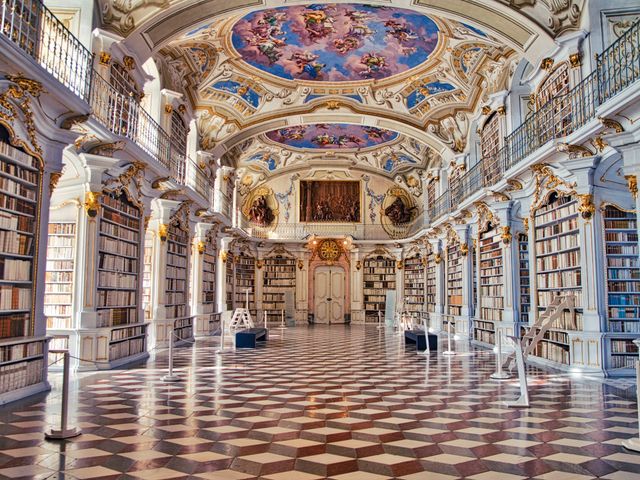 A grand library with murals on the ceilings, marble floors, gilded accents, and walls lined with books.