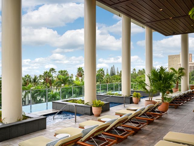 A terrace infinity pool framed by tall columns with seating under the shade of a roof, overlooking views of Waikiki Beach.