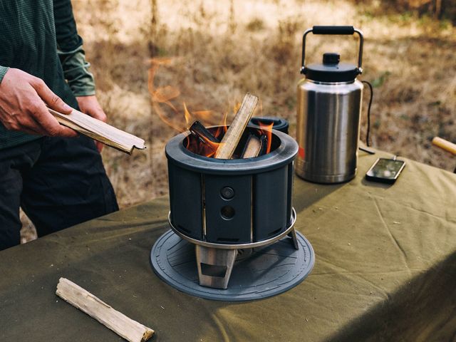 A small fire pit is on a table with flames and wood visible inside, as hands wait to add more wood.