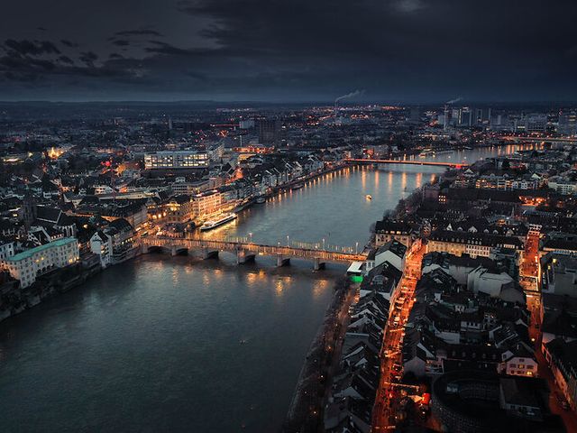 An aerial view of Basel, Switzerland at night with lit-up buildings and a bridge crossing the river.