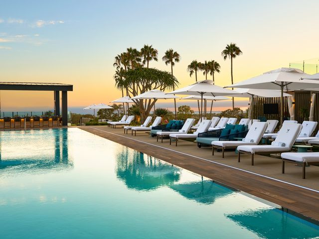 An infinity edge pool lined with white loungers and a bar to the side at sunset, at Vea Newport Beach.