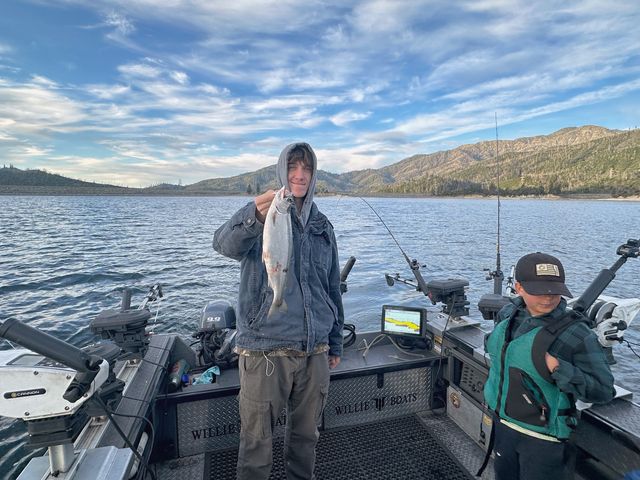 A teenager on a boat holds up a fish just caught while wearing his hood and looking at the camera.