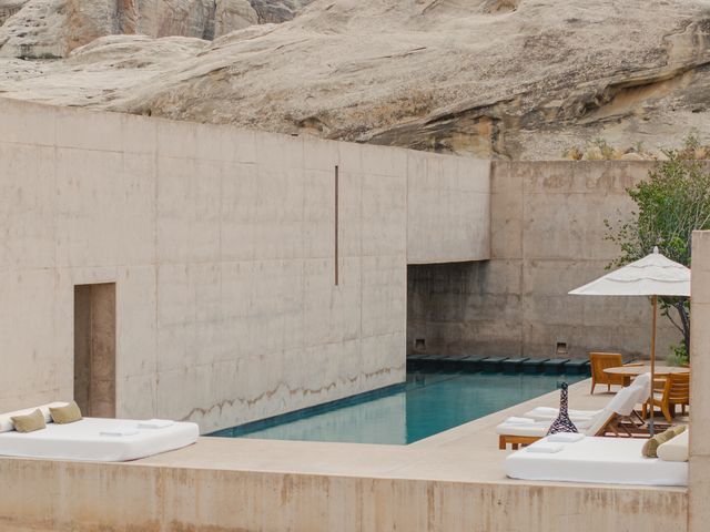 A white stone building built up against a desert mountain wall with an outdoor patio that has a pool and day beds lining the terrace.