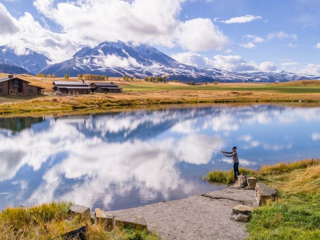 A man stands and fly fishes along a river with a dramatic mountain landscape behind him in Montana.