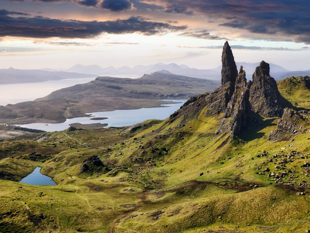 Large rock formations next to grassy areas and views over waterfront land formations on the Isle of Skye under clouds.