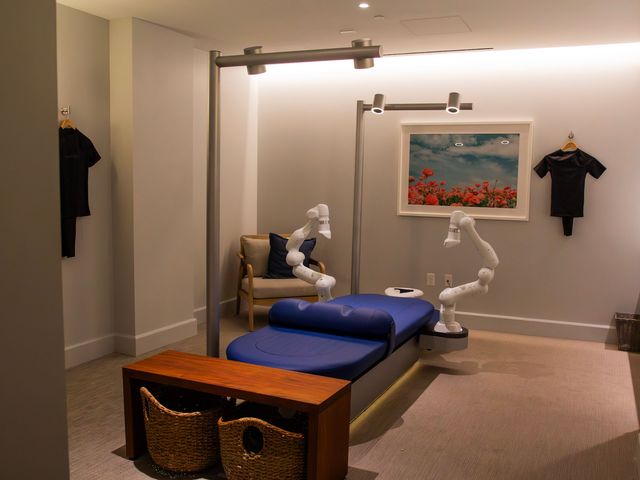An Aescape massage table at The Seabird Ocean Resort & Spa with a blue table, white robotic arms, and a table and chairs in the room and art.