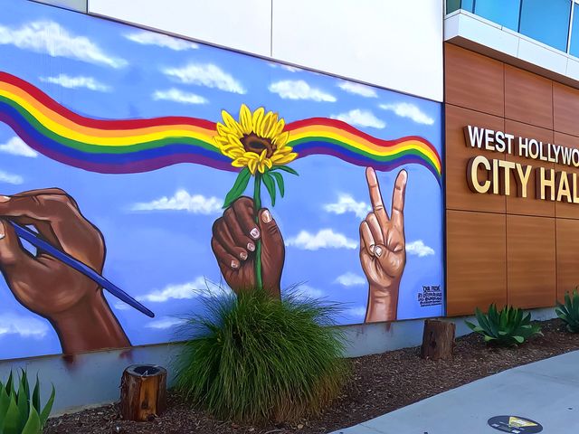 A rainbow painting with three hands on a wall next to a sign for the West Hollywood City Hall.