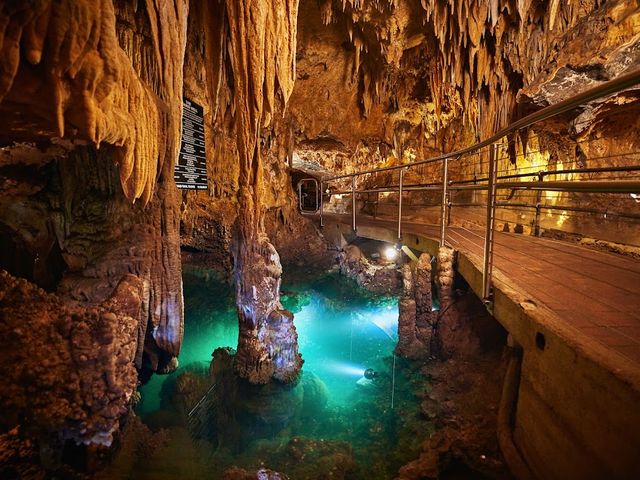 A subterranean area of Luray Caverns with stalactite formations and a walkway positioned over a small pool of water glowing blue.