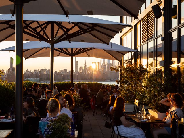 An outdoor patio filled with people sitting at tables under large umbrellas with direct views of the NYC skyline at sunset.