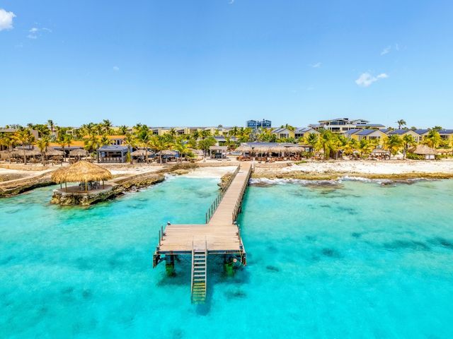 A view of a hotel as seen from the water with thatched huts and long docks reaching into turquoise water.