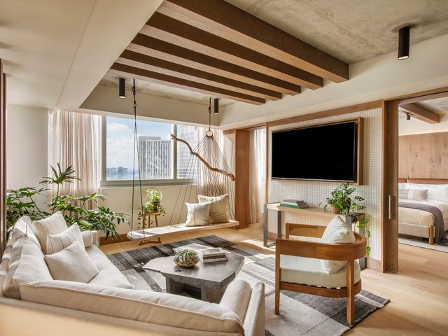 A suite at 1 Hotel Nashville with neutral colors, city views out the window, wooden beams, cream seating, and a bed visible through a doorway.