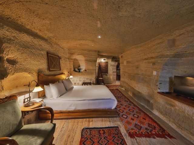 A hotel bed in a cave room carved out of stone with colorful rug on the floor, alcove nooks, lamps, and a chair. 