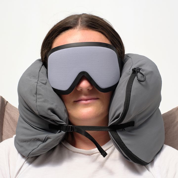 A woman wearing an eye sleep mask with a travel pillow wrapped around her neck.