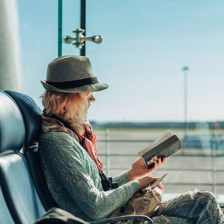 Female traveler wearing hat, scarf, sweater, and jeans reads a paperback at an airport gate.