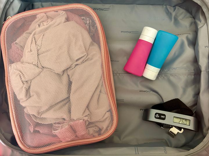 A suitcase half is filled with just a packing cube, blue and pink toiletry bottles, and a digital luggage scale.