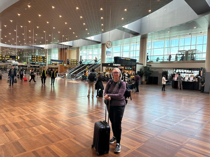 A woman stands in a crowded airport with her hands on her rolling bag wearing a long sleeve shirt and jogger pants.