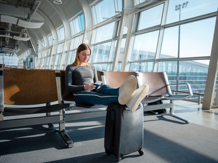 Blonde woman with documents and luggage sitting in the empty airport terminal hall while waiting her flight.