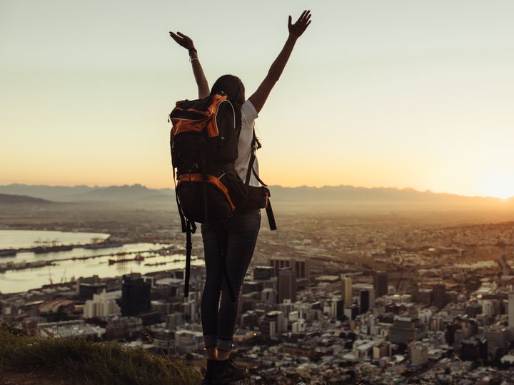 A woman standing on top of a mountain alone wearing a backpack with her hands raised in the air, looking over the city below at sunrise.