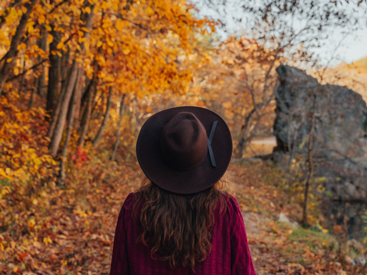 A woman wearing a hat is seen from behind standing in an area amid colorful fall foliage on either side.