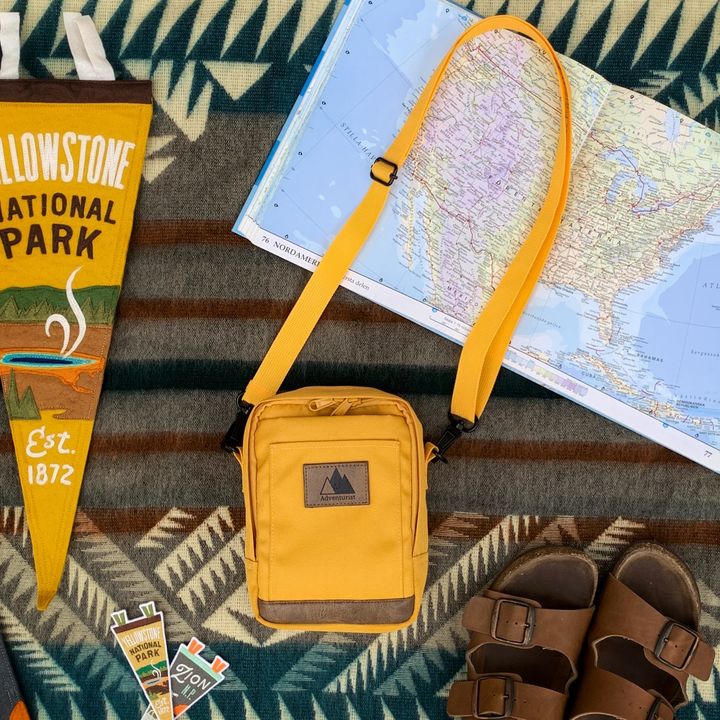 An overhead shot of a yellow Adventurist Backpacks crossbody bag artfully arranged next to a map, flag, and sandals on a tribal print rug.
