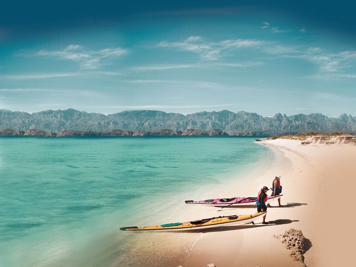 Two people drag their kayaks out of the water on a beautiful beach that has no crowds, white sand, turquoise water, and mountains in the distance. 