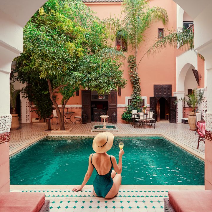 A woman wearing a hat and holding a glass of wine next to an interior Moroccan-style courtyard pool.