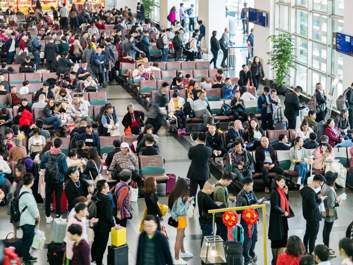 Large crowds of people wait in an airport, as seen from above.