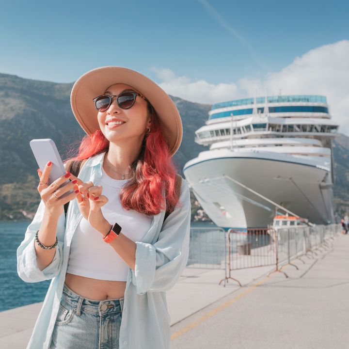 A woman with pink hair wearing sunglasses and a hat on her smart phone and smiling next to a cruise ship.