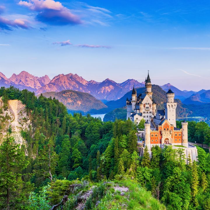The famous Neuschwanstein Castle in Bavaria, Germany, surrounded by rolling hills and mountains, as well as green trees and a lake in the distance.