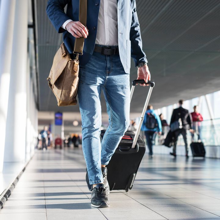 A man with a shoulder bag and hand luggage walking in airport terminal without his face visible.