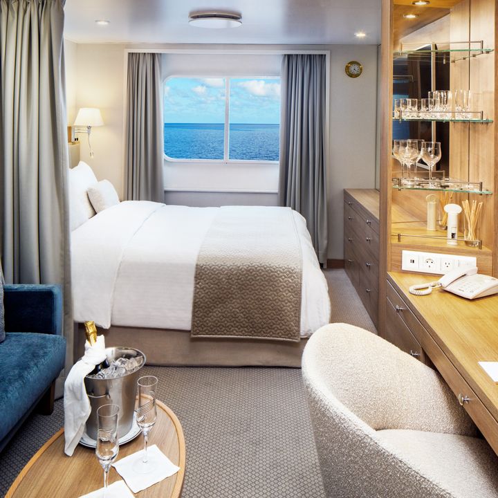 A cruise cabin with a bed and a curtain separating a living area with a couch, table, and desk, in a windowed stateroom.