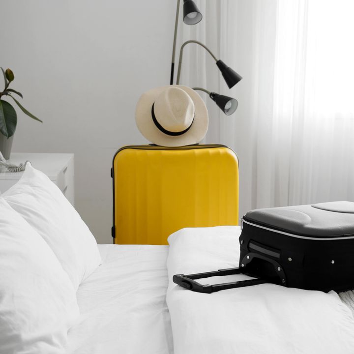 A black suitcase rests on a bed with white linens in a white room, and a standing yellow suitcase with a hat on top stands next to the bed.
