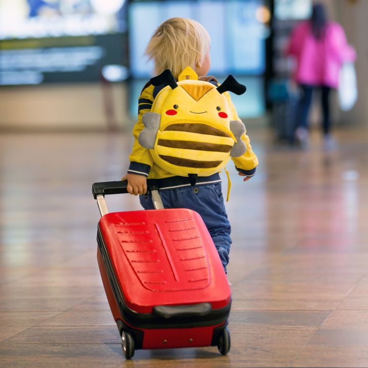 A child pulling a small red suitcase while wearing a bee backpack.