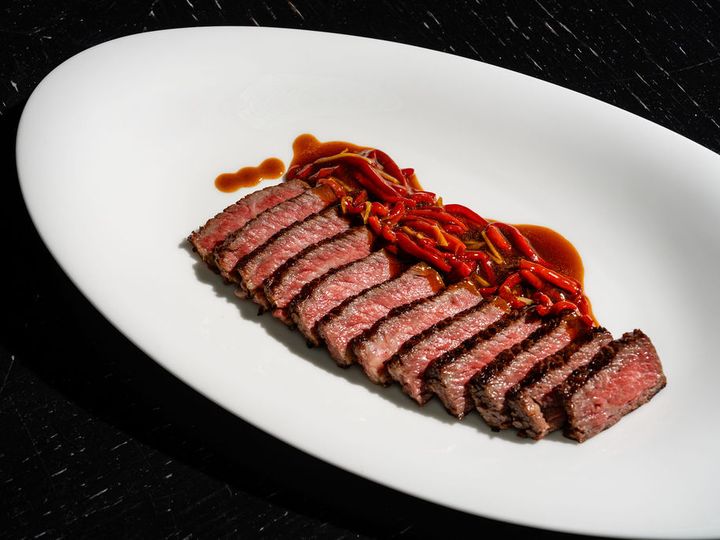 Neatly sliced pieces of medium rare steak arranged on a white plate next to red peppers in a brown sauce.