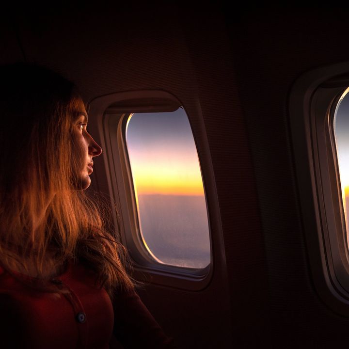 A woman rests her head against the airplane window while looking out at the sunrise.