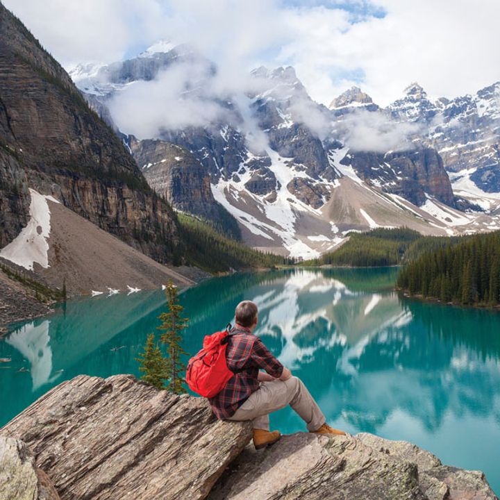 A man with a backpack sits on a rock overlooking snowcapped mountains and a blue lake.