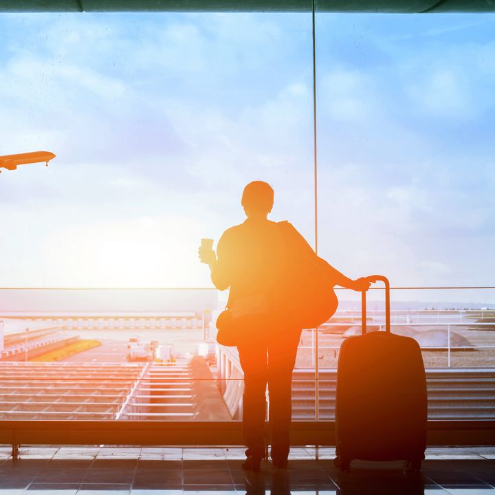 A person is seen looking through an airport window holding luggage and a coffee as light shines through.