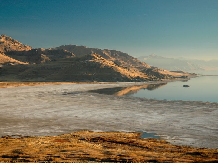 The dramatic landscapes of Antelope Island inside Great Salt Lake with large mountains and a sparse shore all lit gold by the sun.