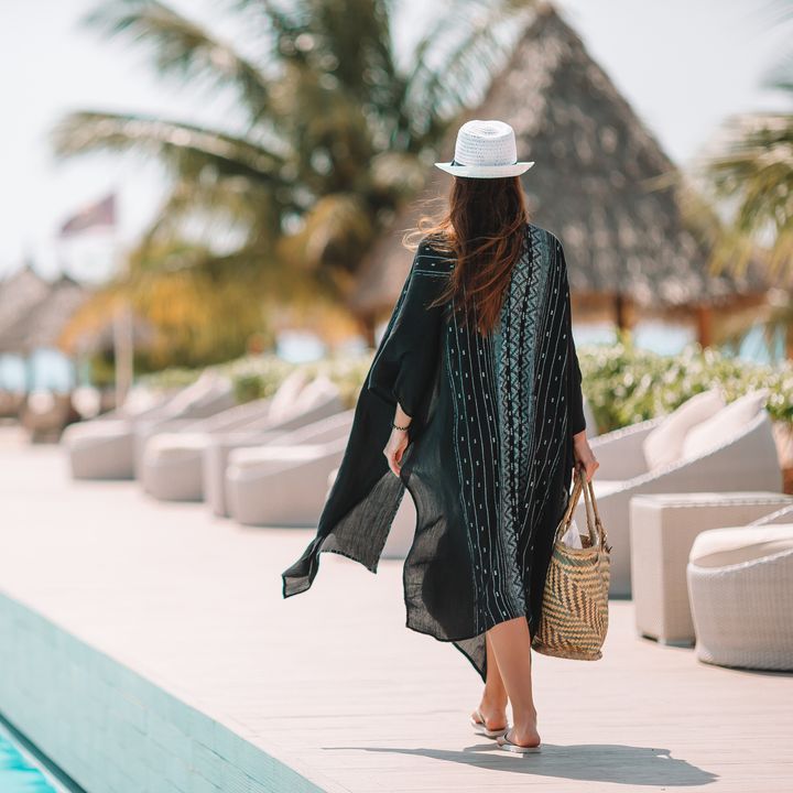 A woman is seen walking by the pool in a caftan and a hat in a luxury hotel resort in a tropical location.