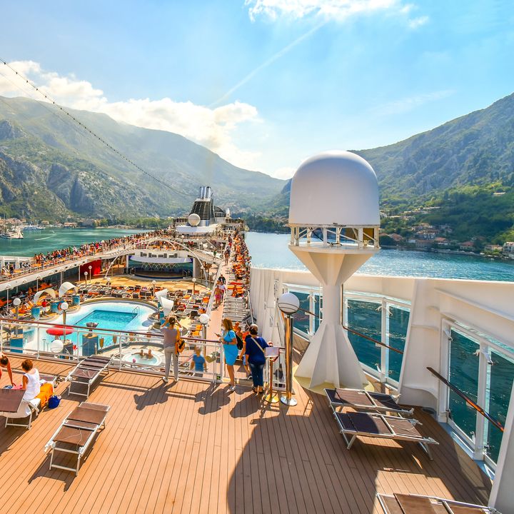 An aerial view over a top deck of a cruise ship with a pool and seating areas as the ship sails in a pretty Mediterranean setting.