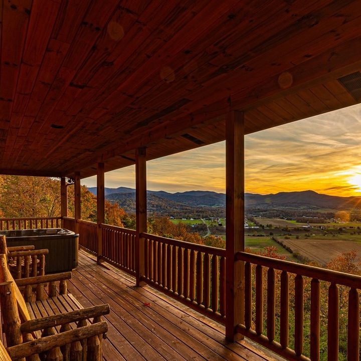 One side of a log cabin with a long wooden deck, two chairs, and views stretching over fields and mountains at sunset in fall.