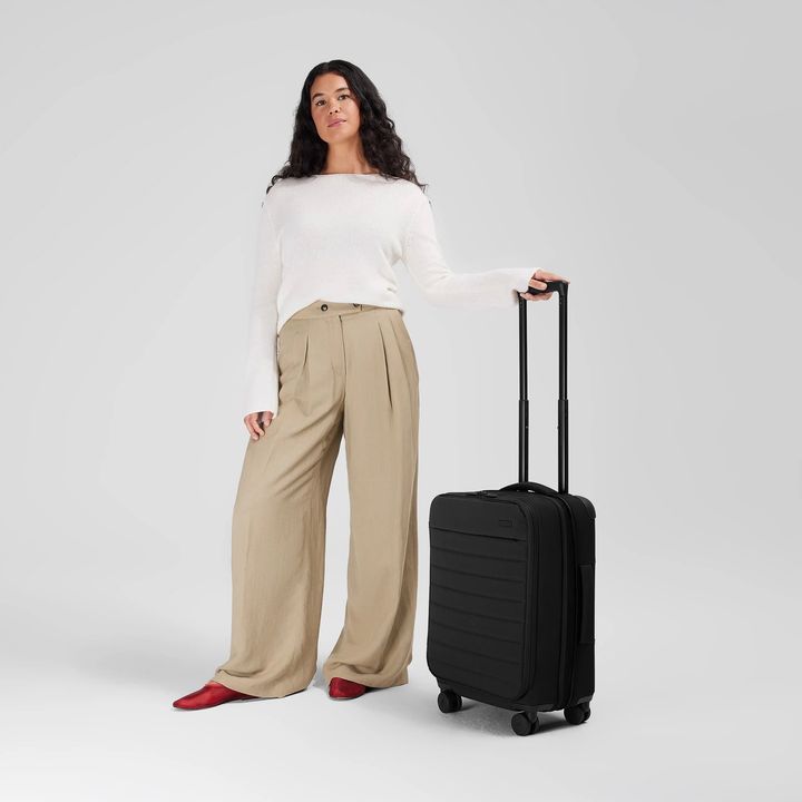 A woman stands against a neutral background holding the handle to an Away wheeled soft-sided carry-on.