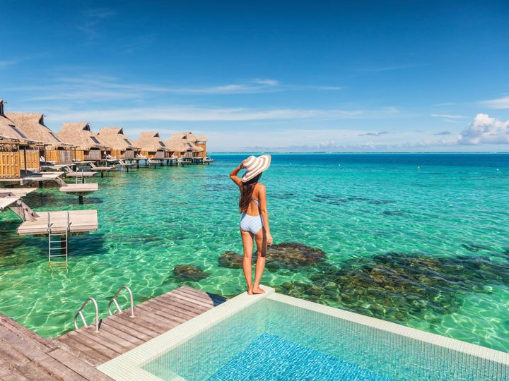 A woman standing next to a pool looking out over the ocean next to a row of overwater bungalows.